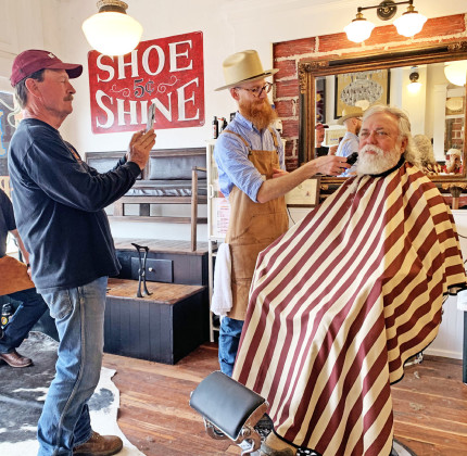 TURKEY BEARD Austen Covin trims Wylie Bomar’s beard Saturday at the historic Ham’s Barber Shop in Turkey, where Bob Wills once gave shaves and haircuts. Snapping a picture is owner Mike Goldsby; taking a break is the shoeshine boy, Josh Kreusel. Shaves, haircuts, shoeshines and tours of the refurbished building were free on Saturday, compliments of Goldsby. Reporter-photographer Hanaba Munn Welch is visible in the bottom right corner of the mirror. | HANABA MUNNWELCH