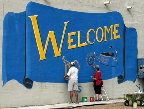 BIG WELCOME was painted on the wall of the Spur Ex-Student Association this summer by renowned artist Vicki Glass Diggs (here, with a little help on July 4 from Daniel Perez).